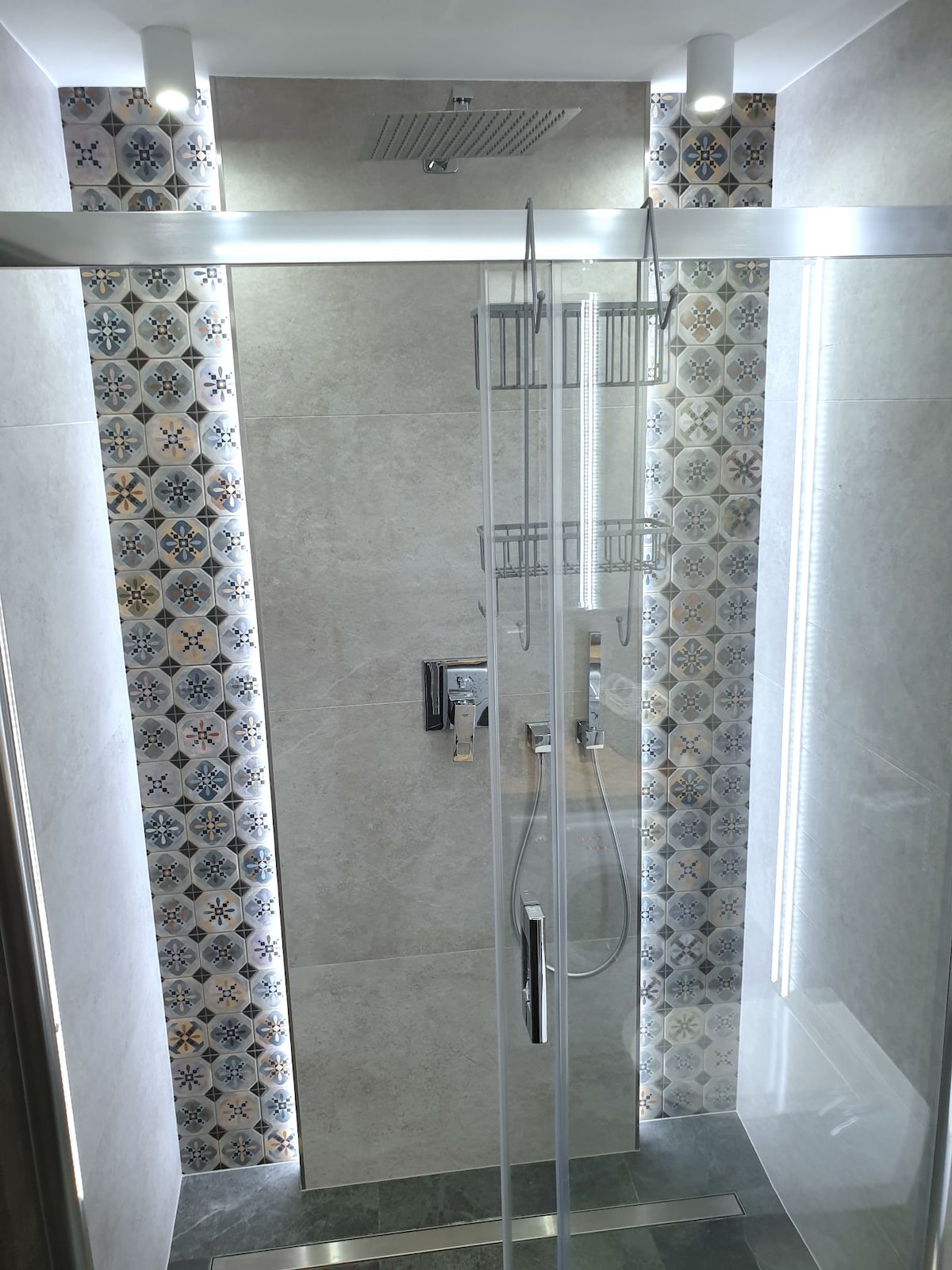 A modern shower area is enclosed with glass panels, featuring intricate patterned tiles along the walls. Soft LED lighting highlights the sleek fixtures and a rainfall showerhead positioned above, emphasizing a clean and functional design.