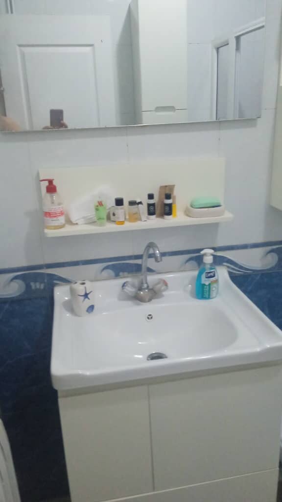 The bathroom features a white sink with a chrome faucet, positioned beneath a mirror. A small shelf holds various toiletries, and blue decorative tiles adorn the wall. The space is compact, with clean lines and a neutral color palette.