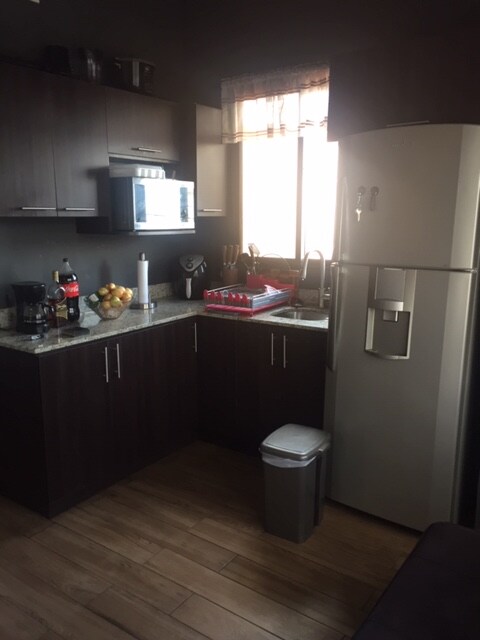 The kitchen features modern dark cabinetry and a light stone countertop. Appliances include a refrigerator, microwave, and coffee maker. A window allows natural light to fill the space, highlighting a bowl of fruit and dishware. The flooring is finished in warm wood tones.