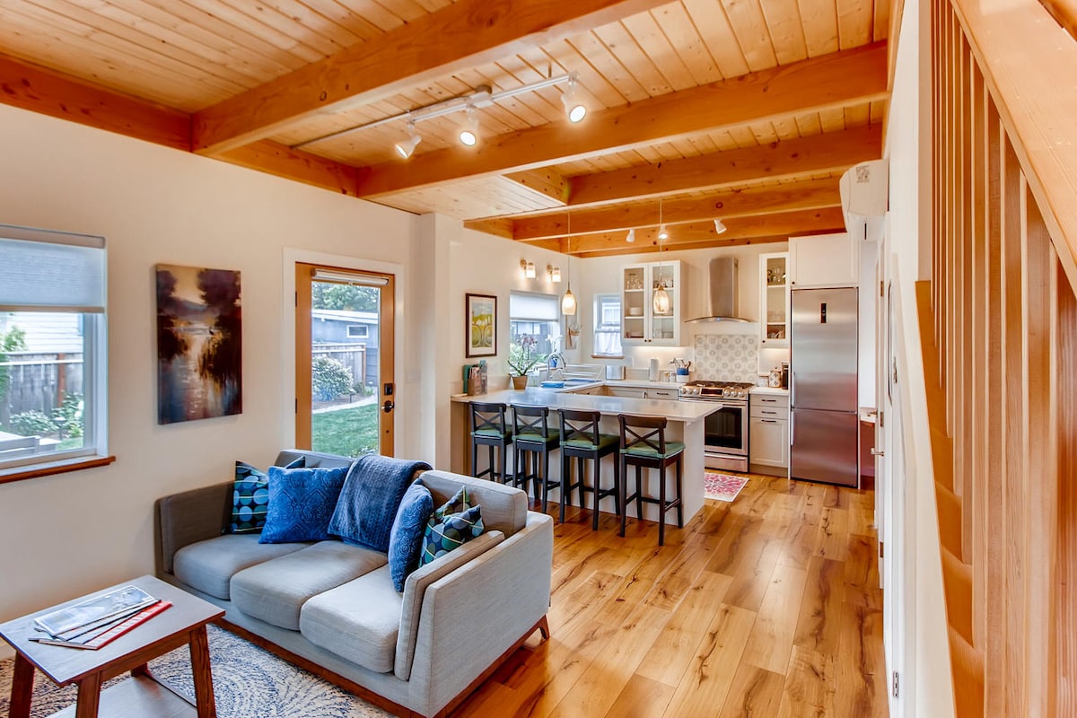 An open living area features a comfortable couch with accent pillows, positioned next to large windows that provide natural light. The adjoining kitchen showcases a modern design with a peninsula and bar stools, surrounded by light wood beams and laminate flooring.
