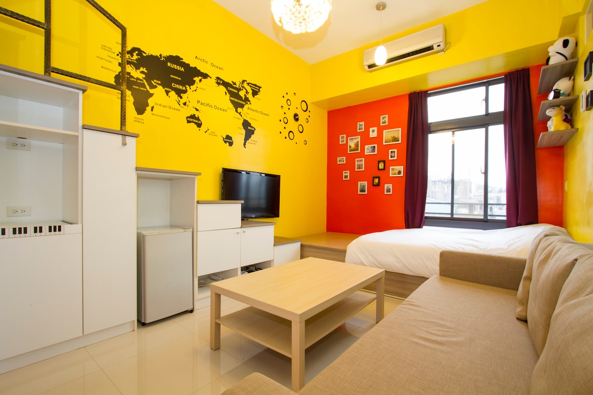 A vibrant living area is adorned with a bright yellow wall displaying a world map. A large window is framed with burgundy curtains, allowing natural light to fill the space. A comfortable sofa faces a low wooden coffee table, with a television and storage units nearby.