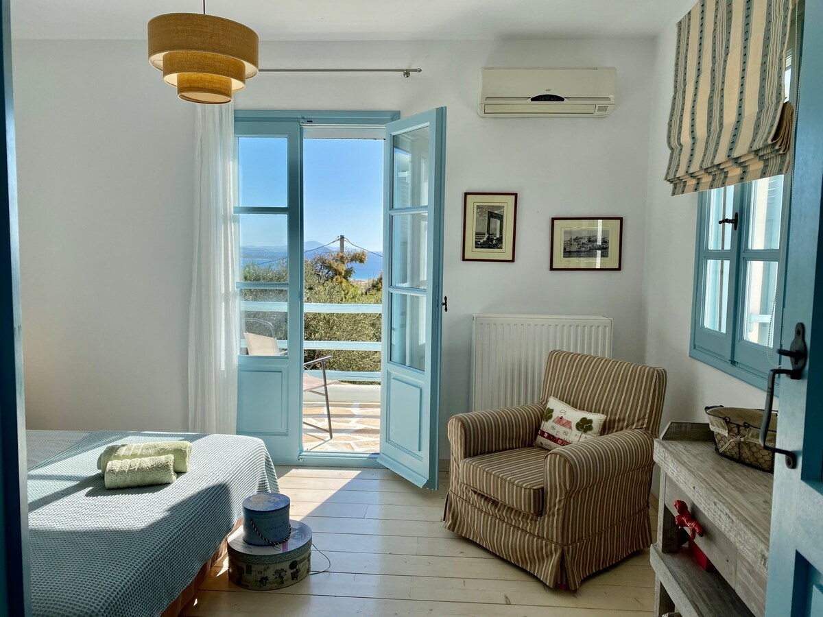 A bright bedroom features a large window with blue shutters, providing views of the sea. A spacious bed is positioned next to a comfortable armchair. Natural light fills the room, highlighting the neutral decor and a subtle accent chair, creating a serene and airy atmosphere.