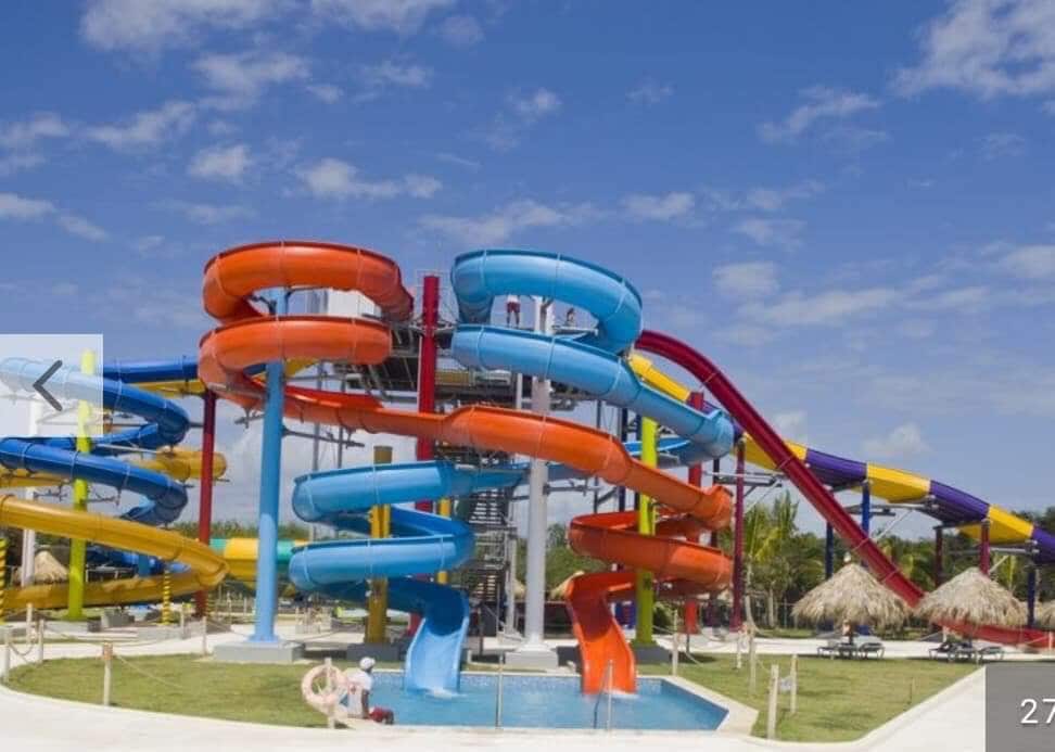A vibrant water park features multiple twisting slides in shades of orange, blue, and purple. A swimming area is located at the base, surrounded by sandy spaces and palm-roofed huts. Clear skies can be seen overhead, enhancing the lively atmosphere.