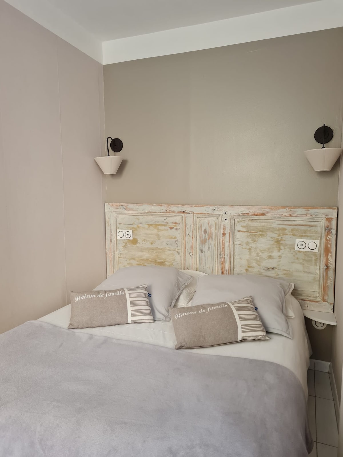 A comfortable sleeping area is defined by a weathered wooden headboard, enhancing the serene atmosphere. Soft gray bedding is layered on the spacious bed, accompanied by decorative pillows. Wall sconces provide gentle illumination, contributing to the soothing palette of muted tones.