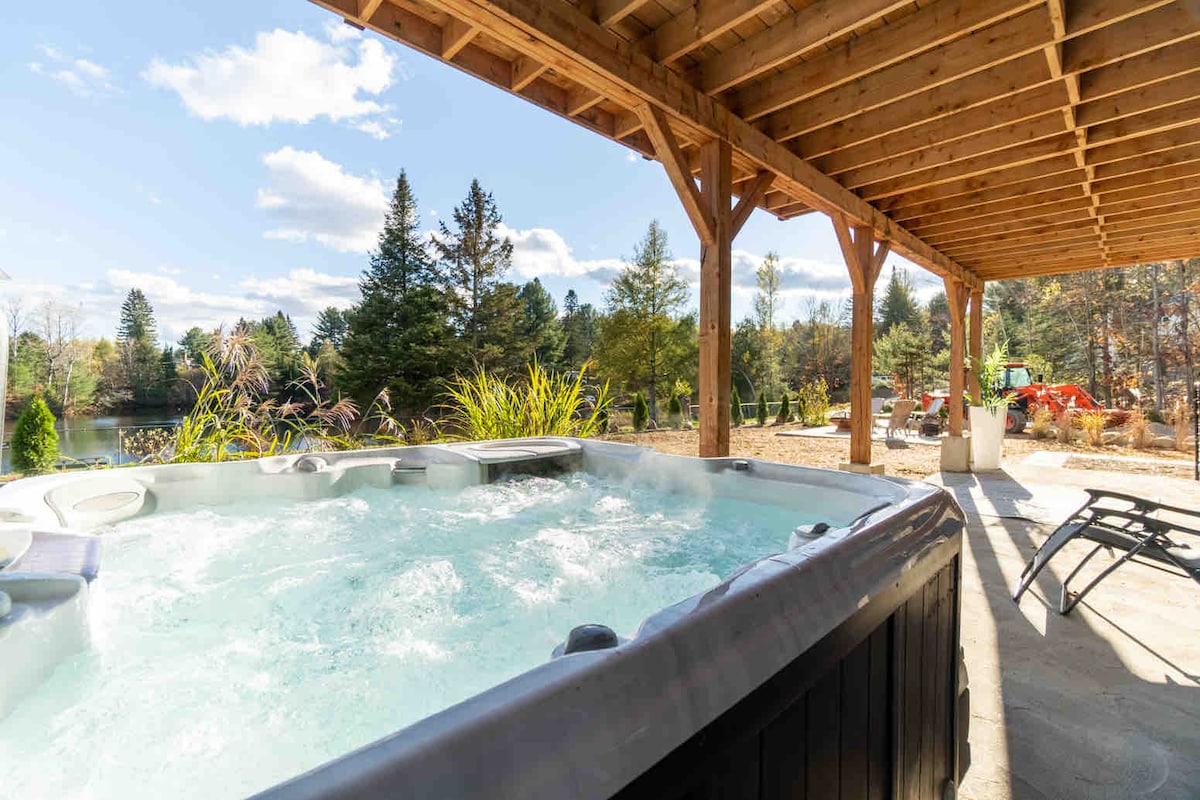 A relaxing outdoor spa is nestled under a wooden structure, providing a perfect view of the surrounding greenery and serene lake. The bubbling hot tub contrasts with the clear blue sky and lush trees, creating a calm atmosphere for unwinding.