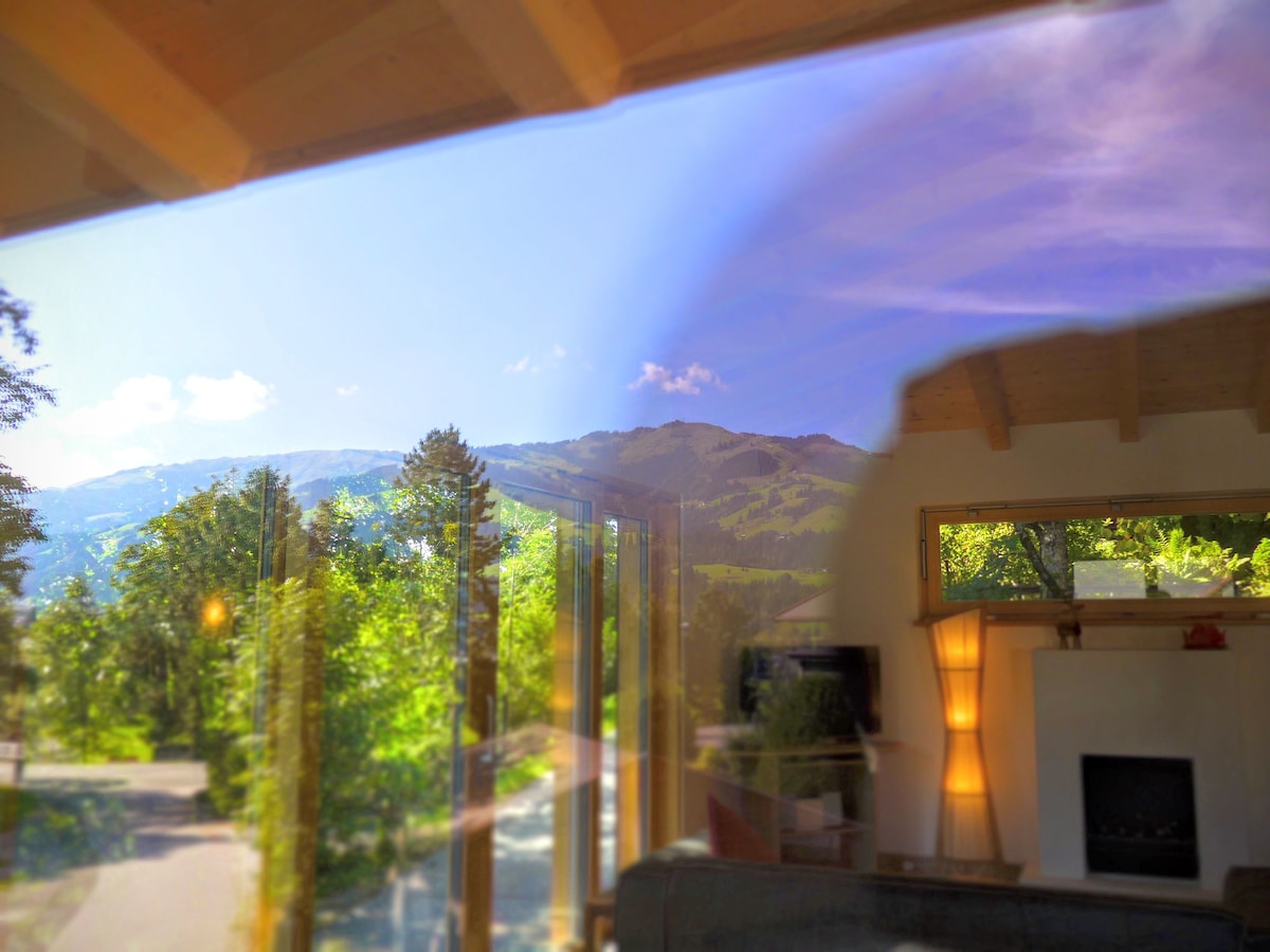 A view from a large window is displayed, showcasing mountains under a clear blue sky. Lush greenery is seen in the foreground, enhancing the connection to nature. The warm light from the sun reflects gently on the glass, creating a serene atmosphere.
