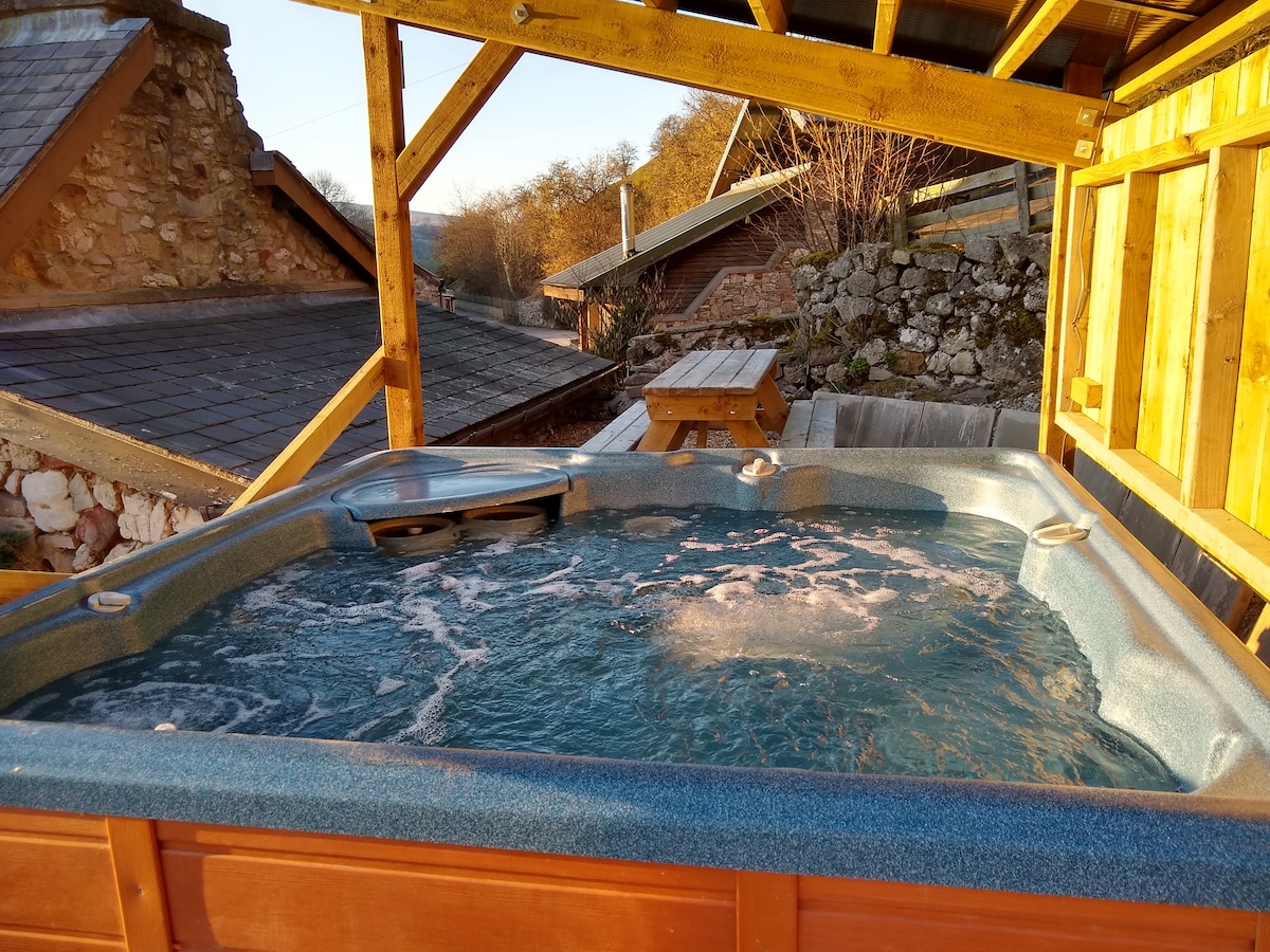 An outdoor hot tub is situated beneath a wooden shelter, featuring a bubbling water surface. Scenic views of the surrounding landscape and rustic structures are visible in the background, creating a serene environment for relaxation.