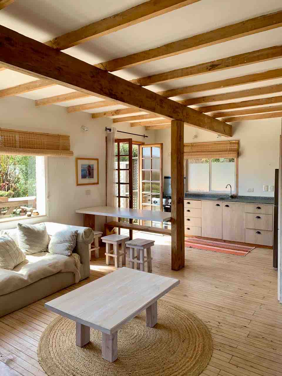 A bright living area features natural wood beams overhead and light streaming through multiple windows. A comfortable sofa is positioned beside a small wooden table, accompanied by two stools. The modern kitchen area includes sleek cabinetry and is adorned with simple decor, creating an inviting ambiance.