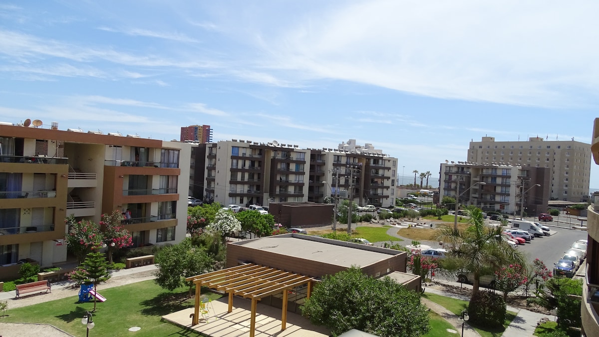 A view of the surrounding buildings from the fourth floor balcony showcases several apartment complexes with balconies and gardens. A landscaped area with trees and benches is visible below, while the distant view includes the sea and sky with scattered clouds.