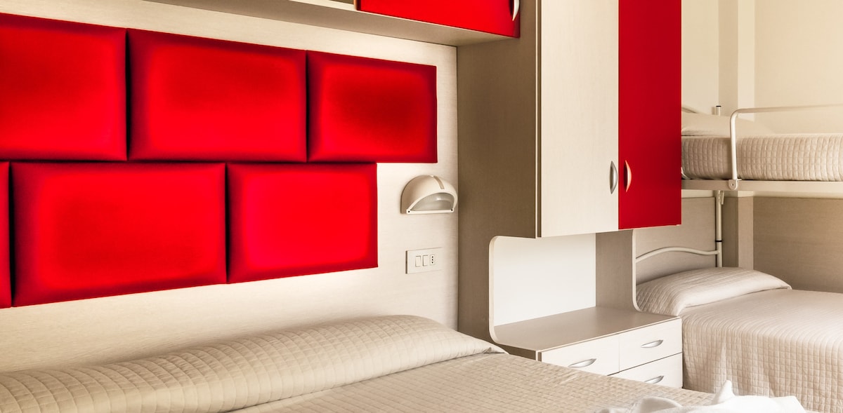 A cozy room is presented, featuring a colorful red wall panel behind the beds. Two spacious beds are complemented by a bunk bed in the corner. A bedside lamp and a built-in cabinet add functionality to the space, with natural light illuminating the interior.