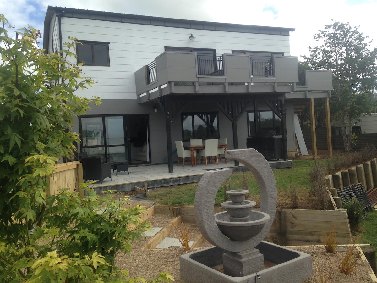 The two-story building features a contemporary design with a large deck above and an outdoor seating area below. A circular water feature is prominently displayed in the landscaped garden, surrounded by gravel and greenery. Large glass doors allow natural light into the unit.