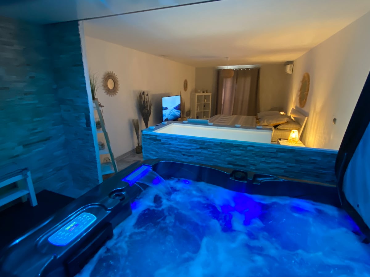 A spacious area features a professional spa setup with bubbling water, illuminated by soft blue lights. In the background, a cozy sleeping space is visible, complete with a large bed and a subtle entertainment unit, creating a serene and relaxing atmosphere.