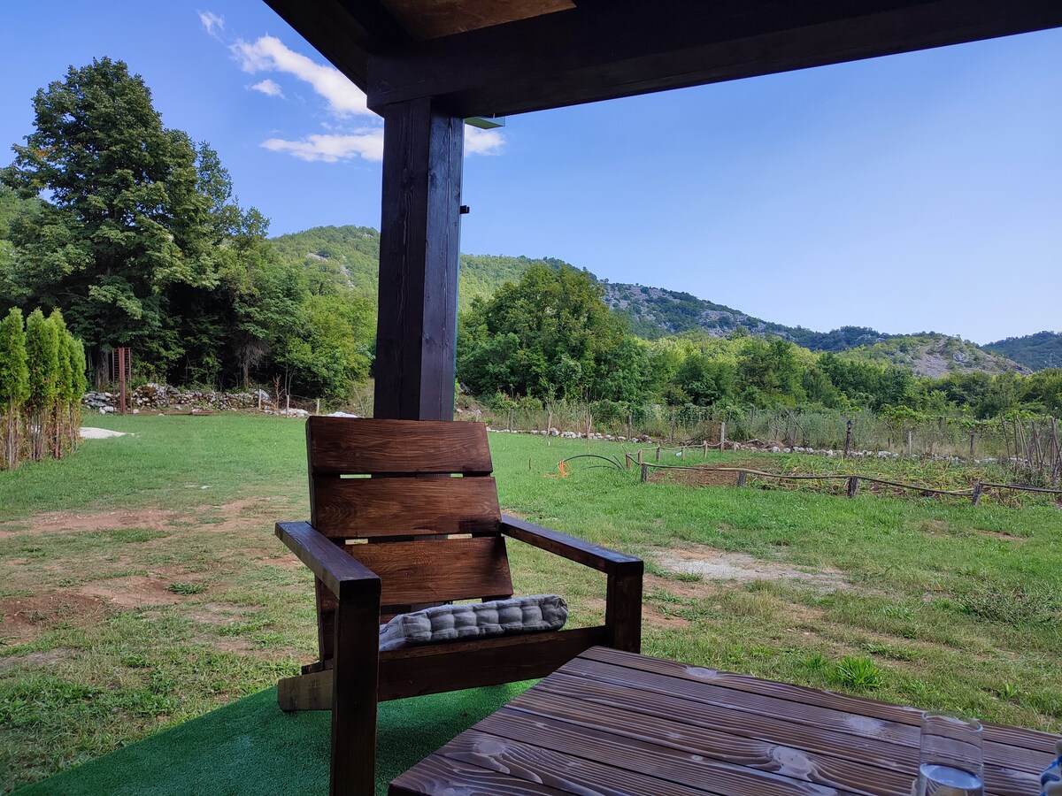 A wooden chair is positioned near a rustic table, overlooking a grassy area. Gently rolling hills and trees can be seen in the background, creating a serene outdoor setting. The sky is clear, with a few clouds visible.