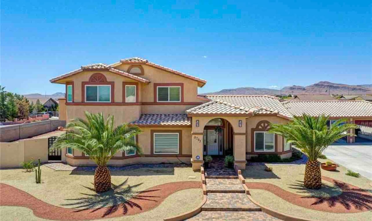 A two-story home with a Mediterranean design features a tiled roof and a welcoming entrance. Palm trees frame the yard, which is laid out in a clean, simple landscape with stone pathways leading to the front door against a backdrop of a clear blue sky.