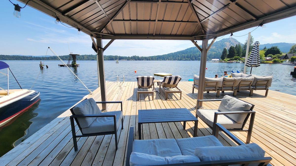 A shaded gazebo accommodates comfortable seating with two lounge chairs and a small table, overlooking the calm lake waters. A boat floats nearby, while the expansive deck offers space for sunbathing and relaxation, framed by scenic views of the surrounding landscape.
