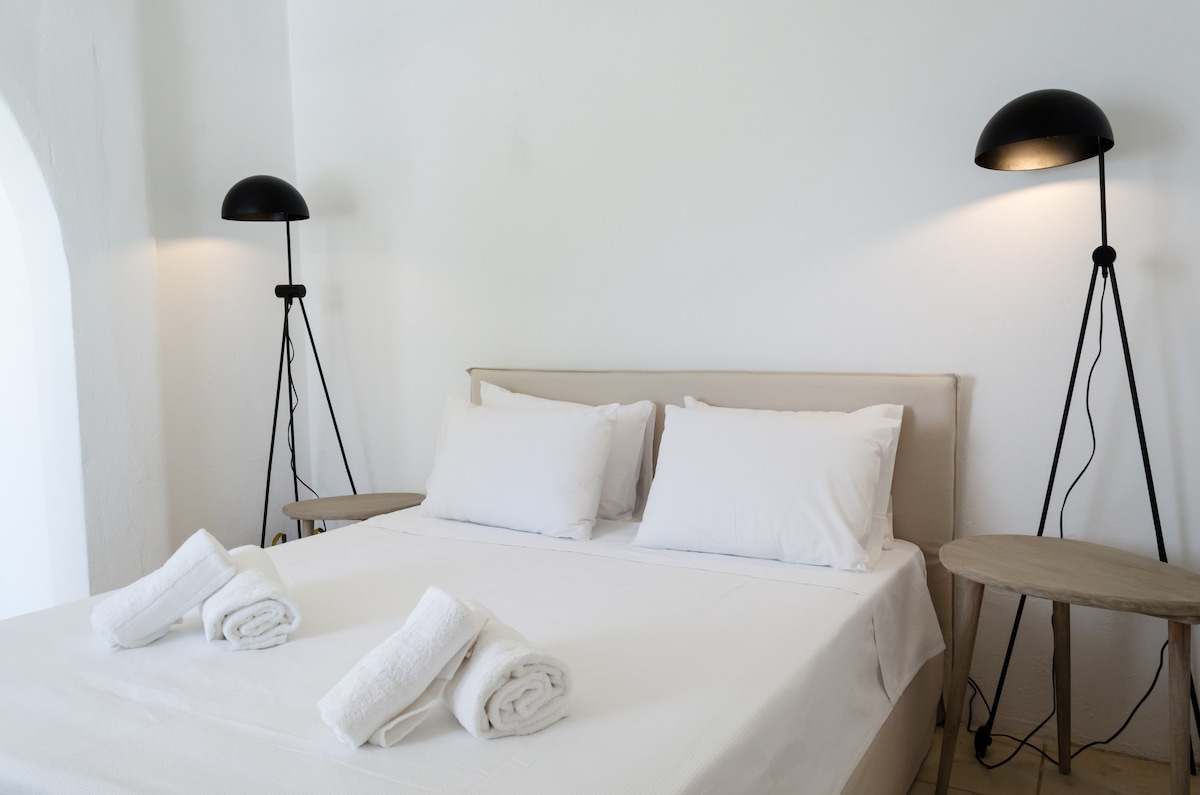 A bright and airy bedroom features a spacious bed with crisp white linens and two neatly arranged towels. Minimalist bedside tables are positioned on either side, complemented by sleek standing lamps that provide soft lighting against the white walls.