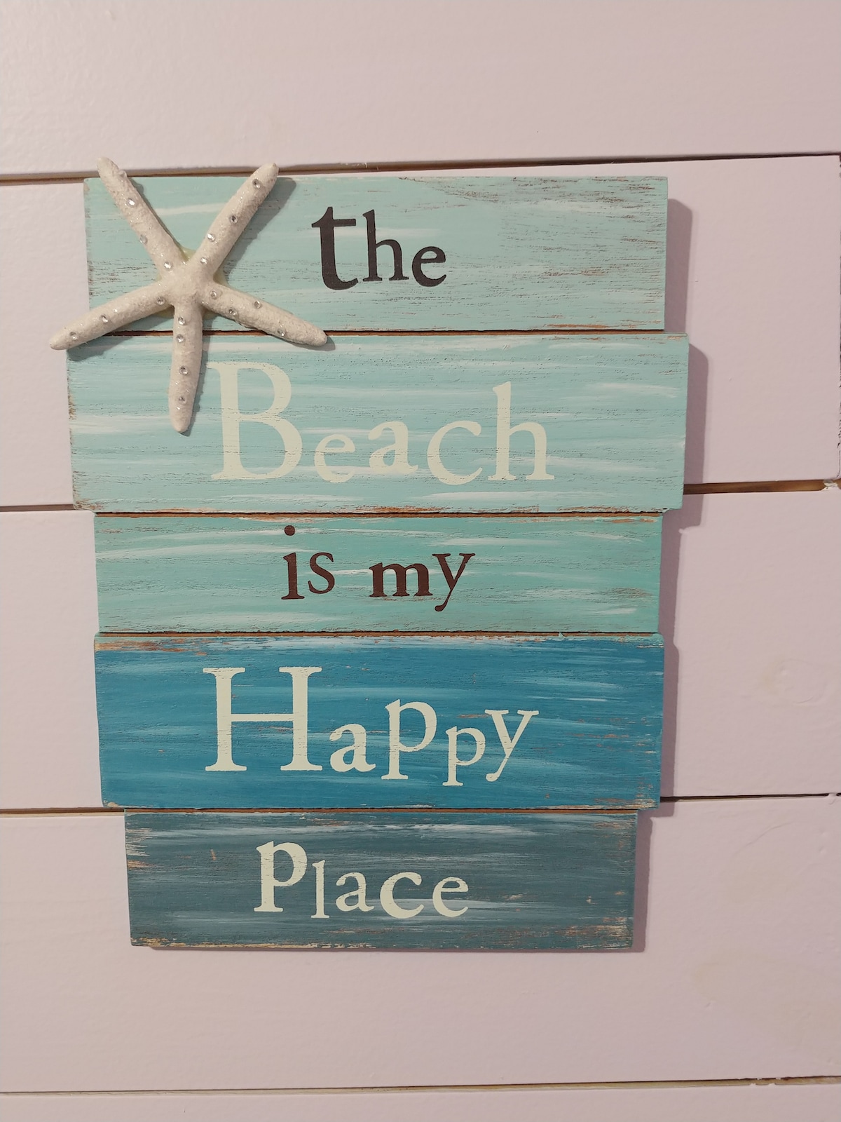 A decorative wooden sign is displayed on the wall, featuring varying shades of blue and text that reads 'the Beach is my Happy Place.' A starfish is attached to the top, adding a coastal touch to the space.