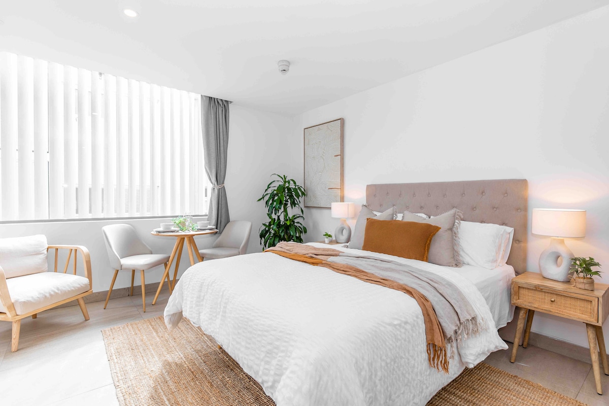 A spacious studio is presented with a large bed dressed in crisp white linens, accompanied by soft brown accents. A small dining table with two chairs sits beside a plant, while warm lighting from bedside lamps enhances a calm ambiance. Vertical blinds allow natural light to filter through.