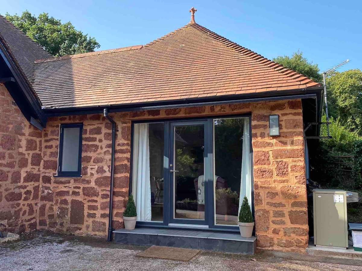 The exterior of the recently converted annexe is displayed, showcasing a charming stone facade with a sloped roof. Tall glass doors provide access to the interior, flanked by two plant pots with small conifer-like plants. Sunlight illuminates the building, highlighting its inviting entrance.