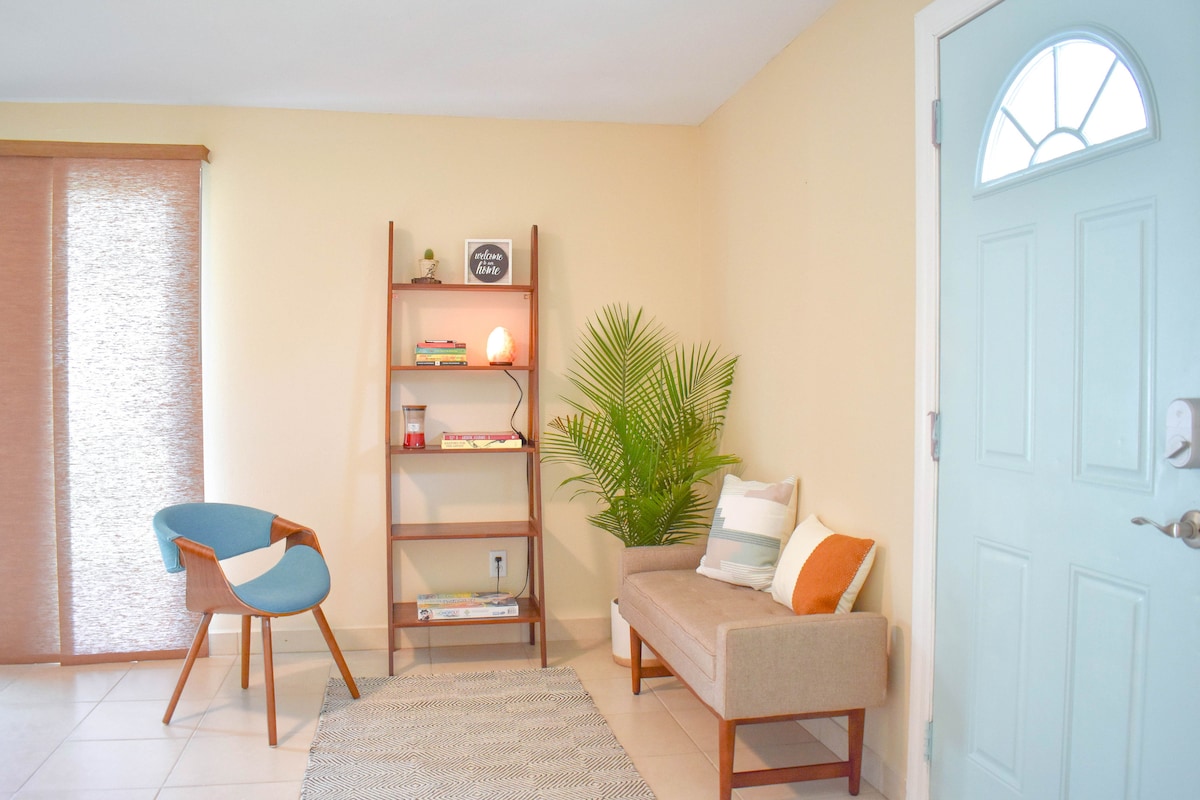 A welcoming entryway features a light blue door and large window, allowing for natural light. A comfortable sofa is paired with a stylish chair and a bookshelf filled with books and decorative elements. A green plant adds a touch of freshness to the space.
