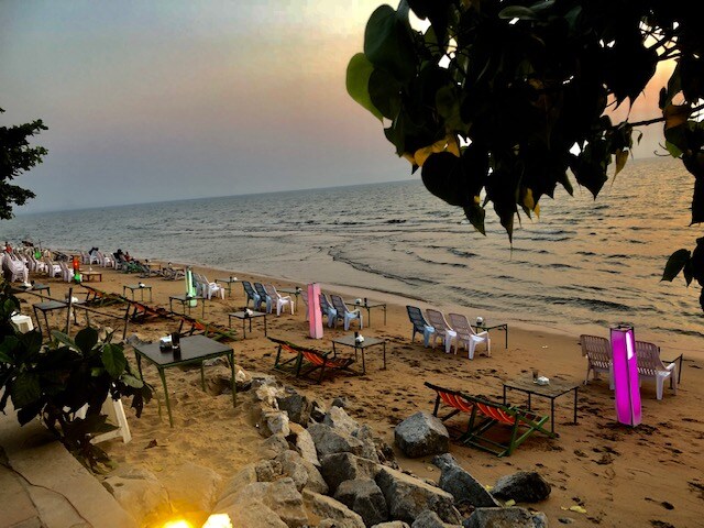 A sandy beach is depicted at sunset, revealing calm ocean waters that enhance the serene atmosphere. Empty loungers and tables are arranged along the shoreline, surrounded by soft hues of pink and orange in the sky. Tropical vegetation is visible at the edge of the frame.