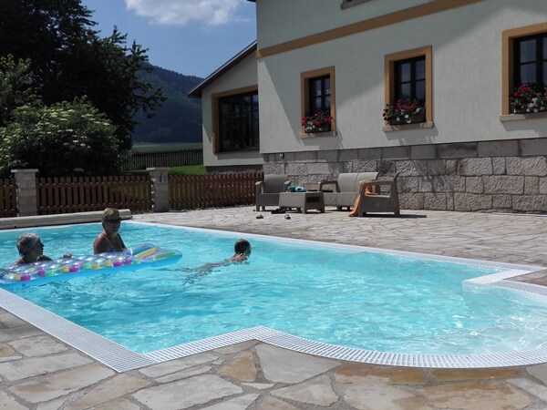 A sparkling pool is surrounded by a spacious patio, featuring lounge chairs and vibrant flower boxes beneath large windows. A child can be seen swimming, while two adults relax on floats, all against a backdrop of greenery and the distant mountains.