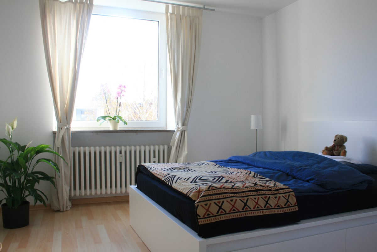 A bright bedroom features a spacious bed covered with a patterned blanket, complemented by a teddy bear resting on the side. A window dressed with light curtains allows natural light to fill the room, alongside a potted plant near the baseboard heater.