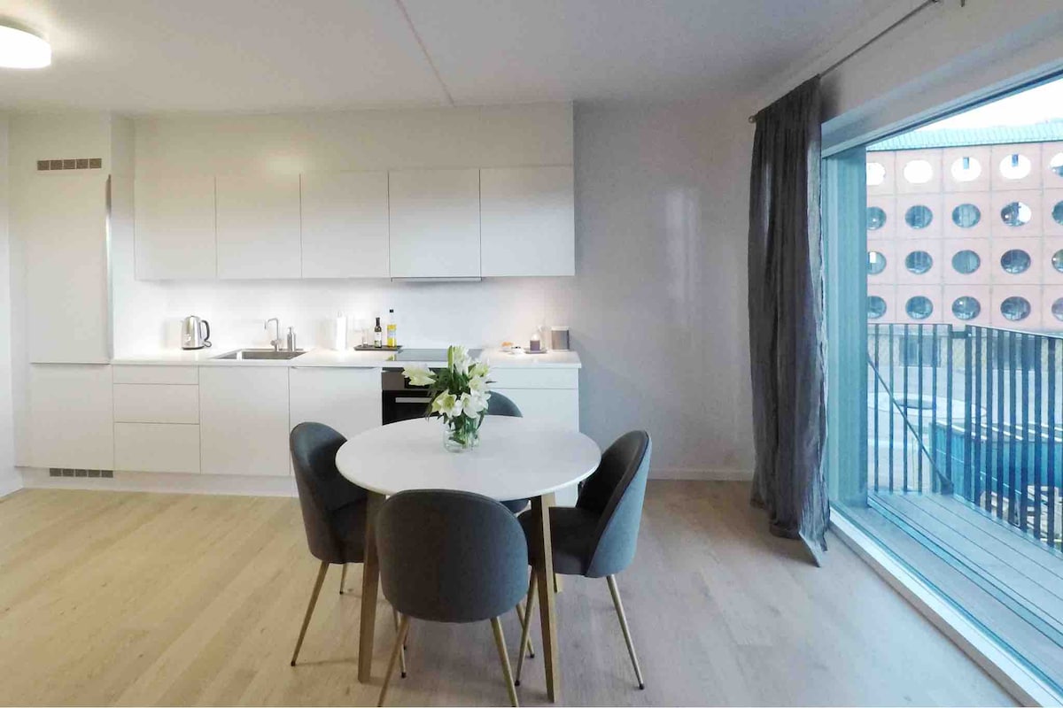 A modern kitchen area is presented, featuring sleek white cabinetry and a round dining table with four upholstered chairs. Natural light fills the space from the large glass door that opens to the balcony, enhancing the minimalist design.