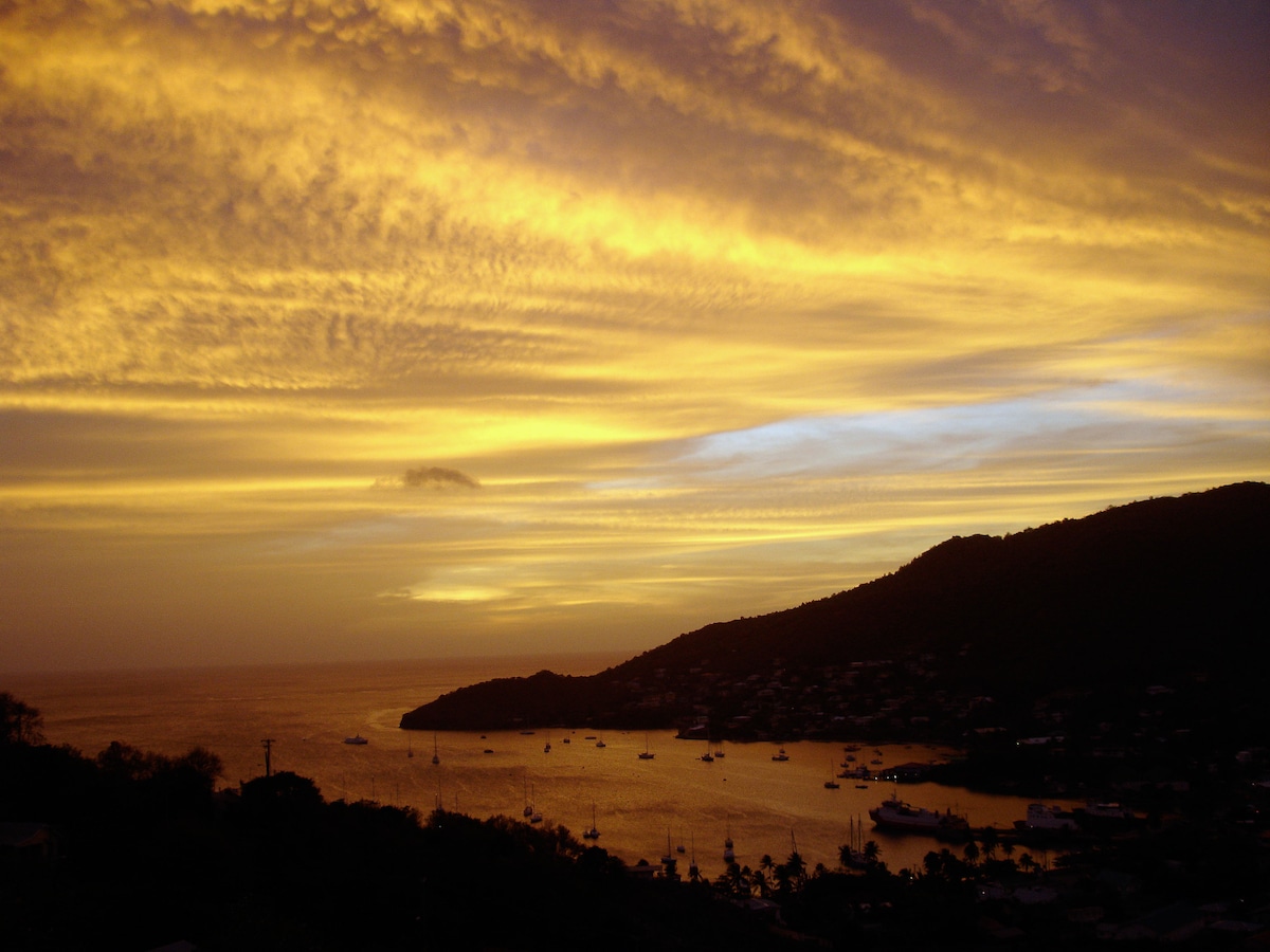 A sunset view over a coastal landscape is captured, displaying vibrant hues of orange and yellow in the sky. The horizon meets a tranquil body of water framed by hills, with silhouettes of boats anchored in the bay, contributing to a serene atmosphere.