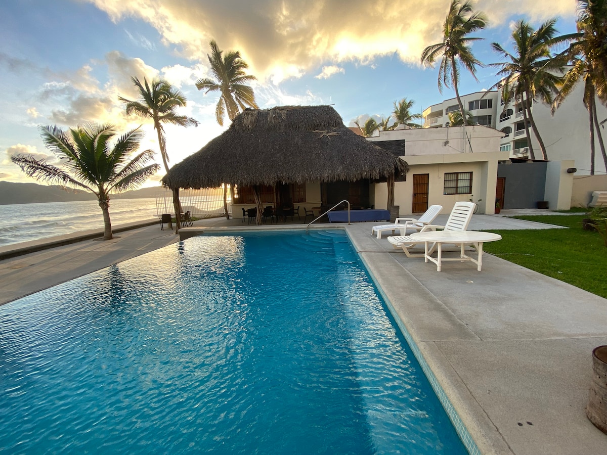 A large pool is situated in front of a traditional palapa, providing shade near the water. Comfortable lounge chairs are placed on the pool deck. The scene is framed by palm trees and a serene view of the beach, with soft clouds in a colorful sky.