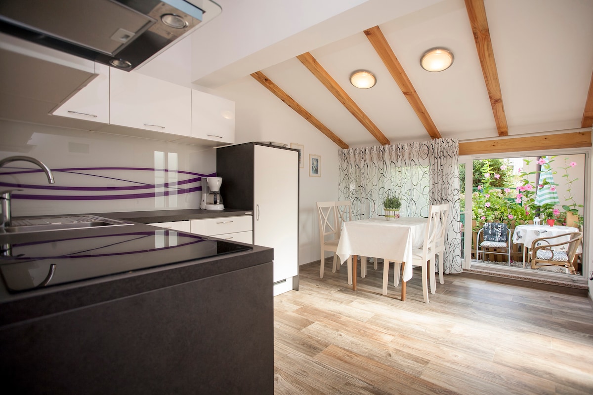 A modern kitchen area is featured with sleek cabinetry and a black countertop, complemented by a coffee maker. A dining space with a white table and chairs is visible, illuminated by natural light from large windows surrounded by greenery.