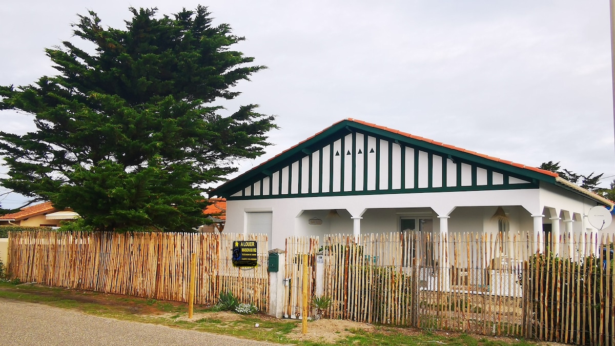 The exterior of the villa showcases a white facade with green trim and a red tile roof. A wooden fence made of vertical planks surrounds the property, with a large cyprus tree nearby. The covered porch is supported by white columns, providing a shaded entry area.