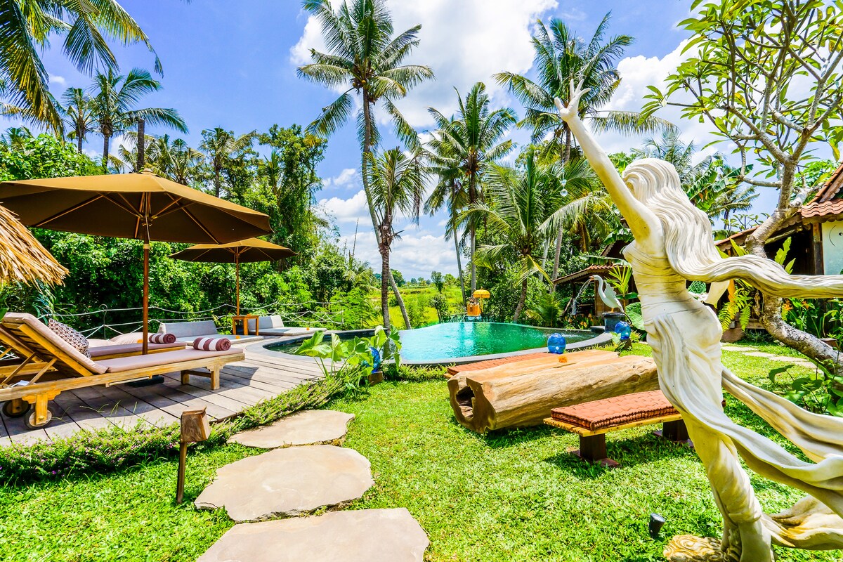 An inviting pool area is surrounded by lush greenery and palm trees. A wooden deck features a sunbed and a shaded seating area. A stone pathway leads through the garden, with a decorative sculpture gracefully positioned in the foreground, creating a serene ambiance.