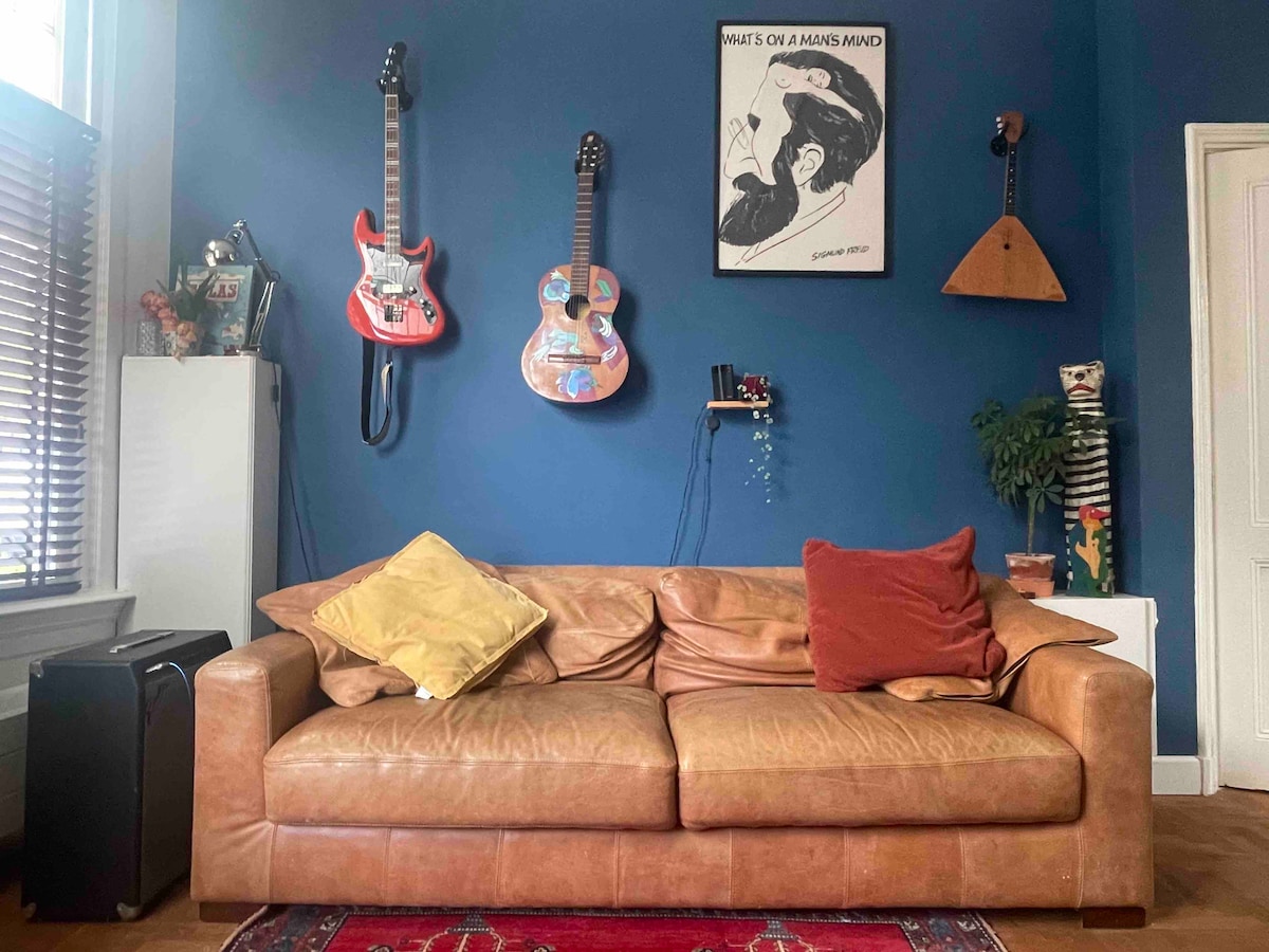 A comfortable sofa is set against a blue wall adorned with guitars and a unique wall art piece. The sofa features brown leather upholstery, complemented by decorative cushions in shades of yellow and red. A small cabinet and a decorative object are also present.