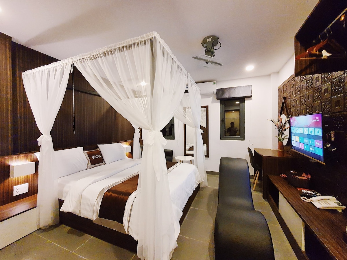 A modern room is presented with a four-poster bed draped in sheer white fabric. Natural light enters through large windows, highlighting a work desk and a television mounted on the wall. Dark wood accents complement the light-colored bedding.