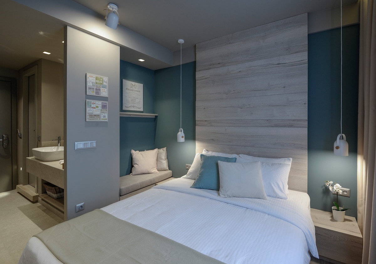 A spacious sleeping area features a queen size bed with light bedding and decorative cushions. Soft lighting highlights the natural wood accent wall. A cozy nook with seating is visible, as well as a modern bathroom entrance in the background.