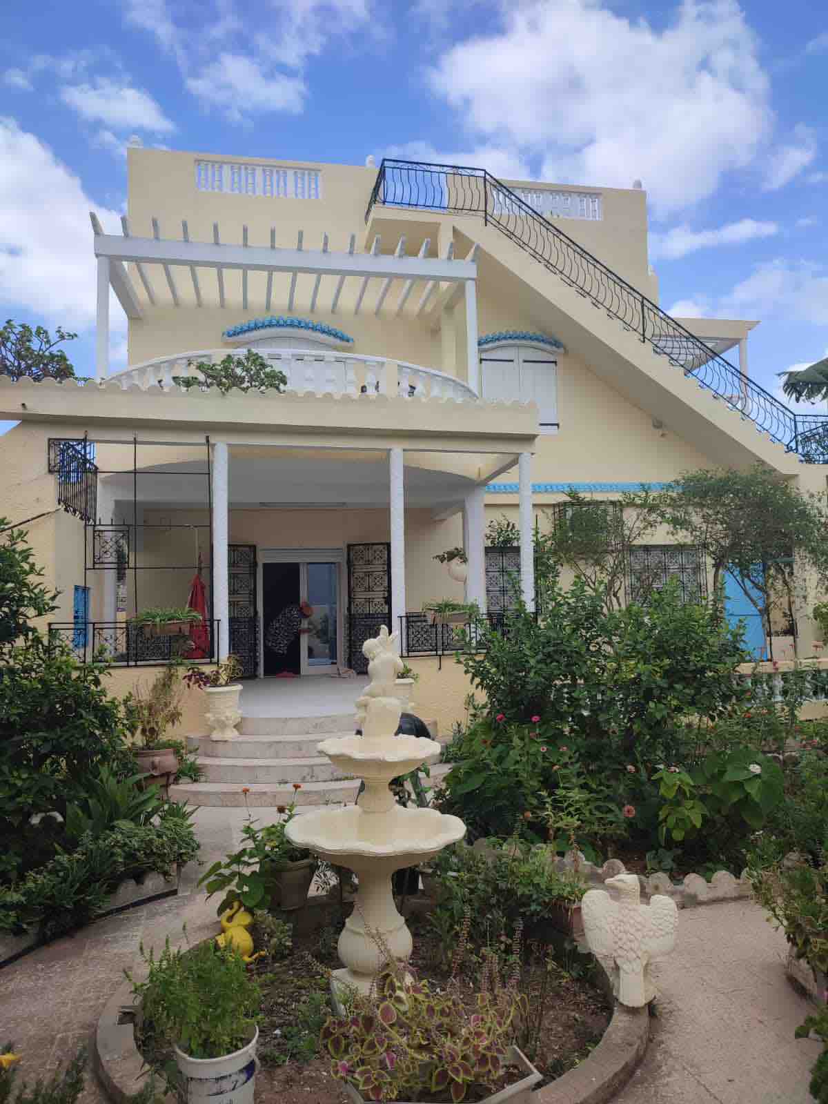 A two-story building with a light yellow facade and blue accents is surrounded by a lush garden. A decorative fountain is centrally located, with vibrant greenery and flowers adorning the outdoor space. A staircase leads to the upper level, enhancing the structure's architectural appeal.