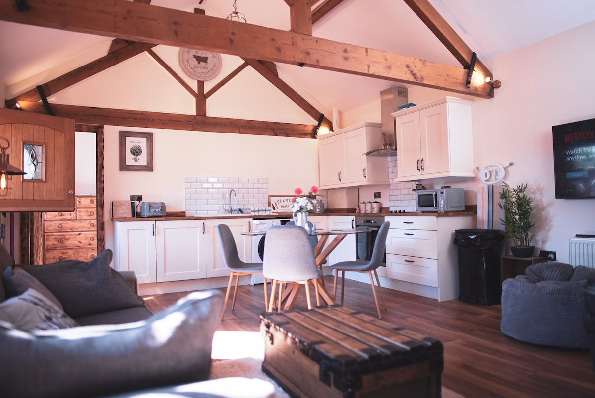 An open-plan living space features wooden beams supporting a high ceiling. A modern kitchen with white cabinetry is complemented by a dining table surrounded by four chairs. Comfortable seating is arranged nearby, with natural light enhancing the warm tones of the wooden flooring.