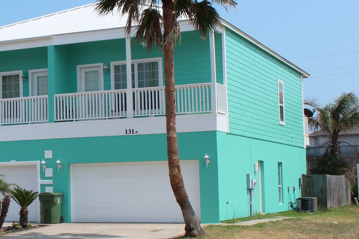 The exterior of a two-story townhouse is depicted, featuring a bright turquoise facade. A palm tree is positioned in front, providing natural greenery. The house includes a covered balcony and a two-car automated garage, showcasing a welcoming beachside aesthetic.