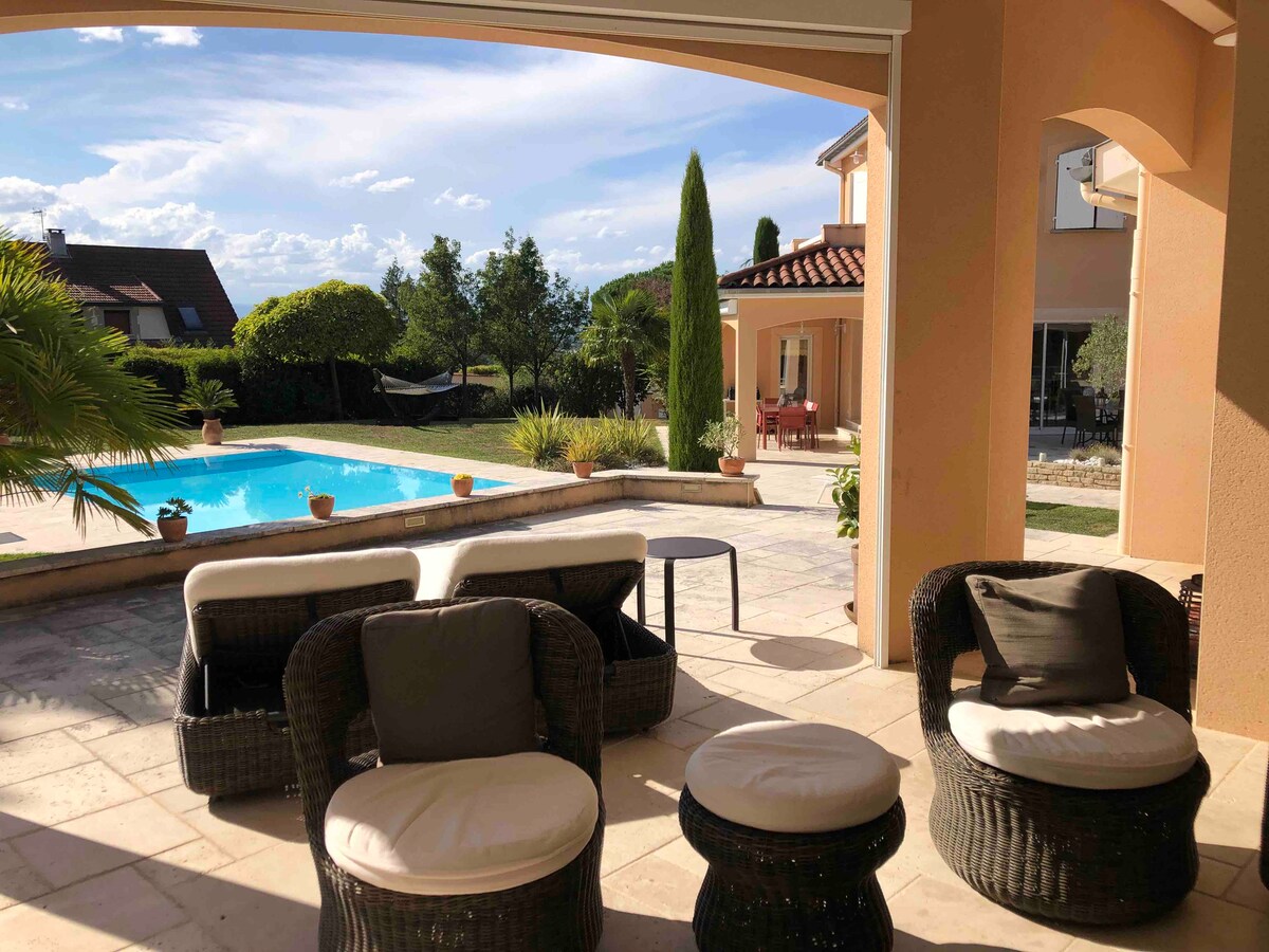 An outdoor patio area provides a view of the pool and surrounding garden. Four wicker chairs are arranged around a low table, with cushions for comfort. Lush greenery and well-maintained shrubs frame the tranquil scene, enhancing the peaceful ambiance of the space.