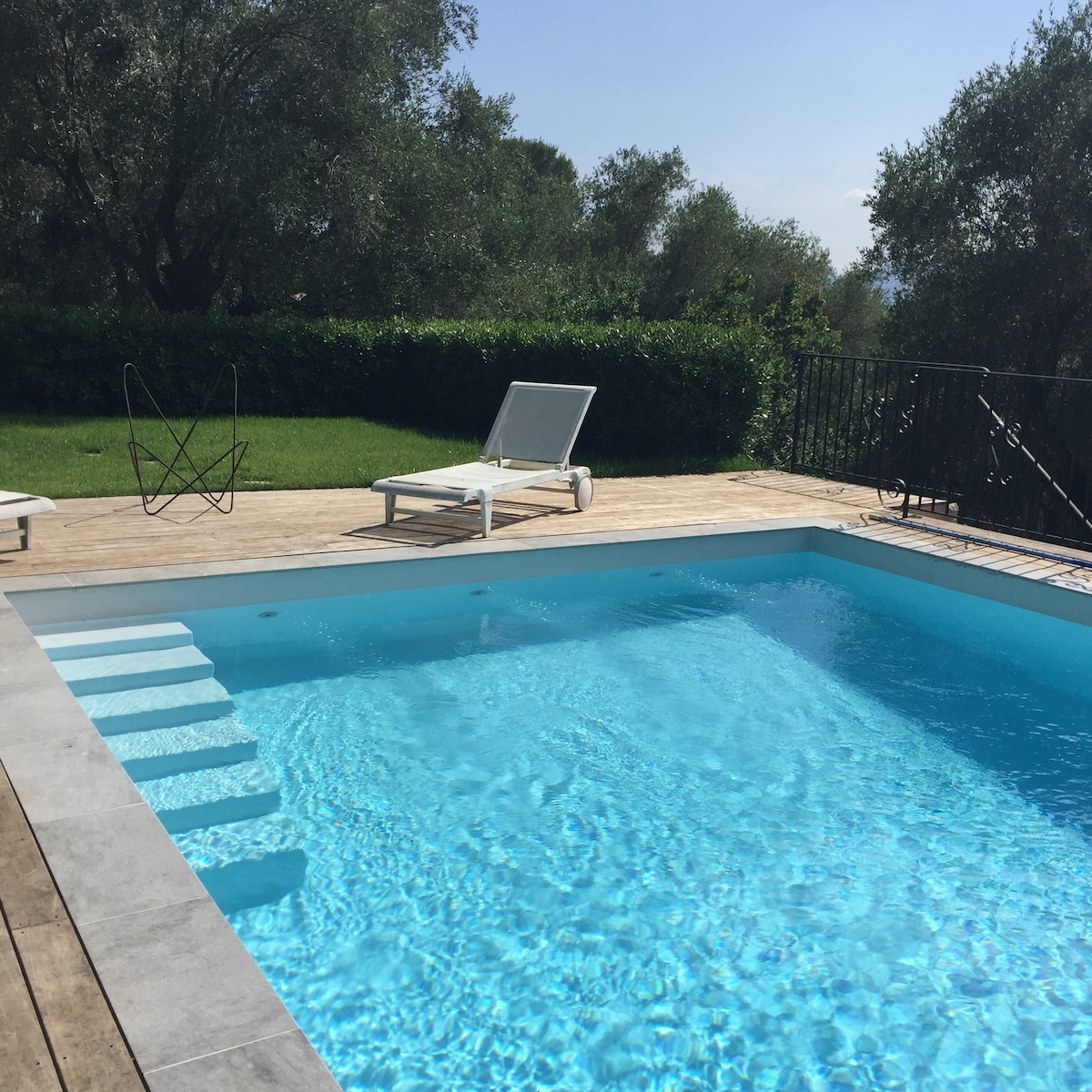 A heated swimming pool is framed by wooden decking and surrounded by lush greenery. The pool features gentle steps leading into the water and is accompanied by a lounge chair and a small side table, offering a serene space for relaxation.