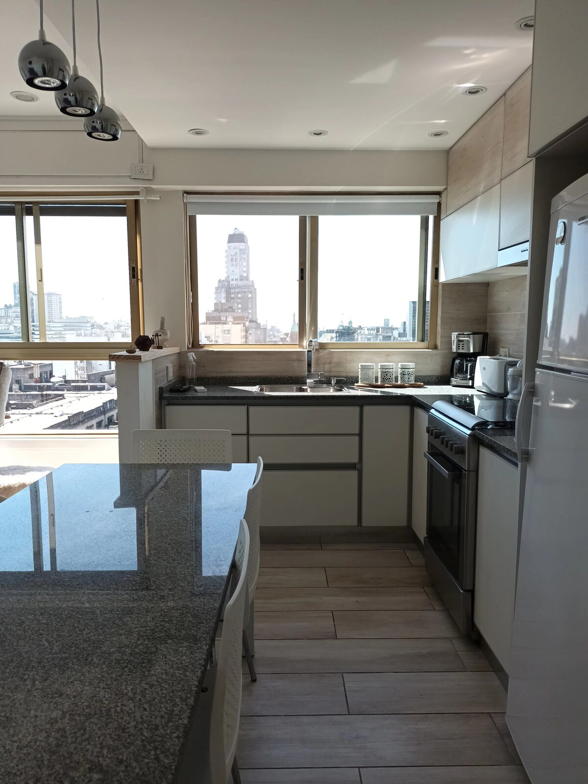A modern kitchen is presented with a granite countertop and stainless steel appliances. Large windows provide abundant natural light and showcase views of the city skyline, including a prominent building. The space includes a coffee maker and dishware arranged neatly on the countertop.
