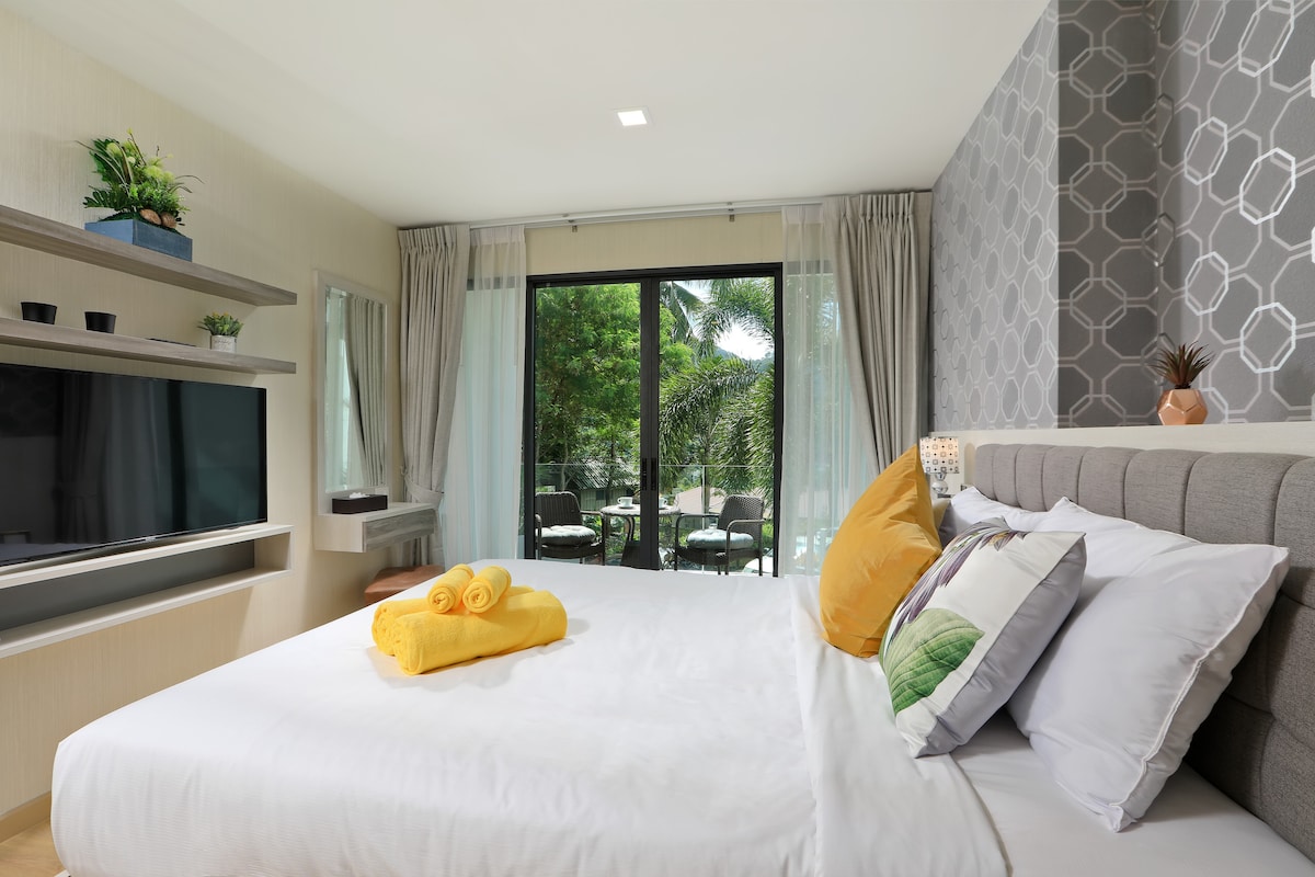 A spacious bedroom features a comfortable bed adorned with bright pillows and neatly rolled towels. Large glass doors lead to a private balcony with views of greenery. A modern shelf and a television are part of the decor, while natural light fills the space.
