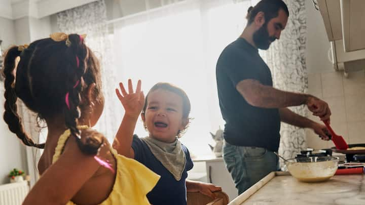 A father makes pancakes while his young son and daughter play next to him at the kitchen counter.