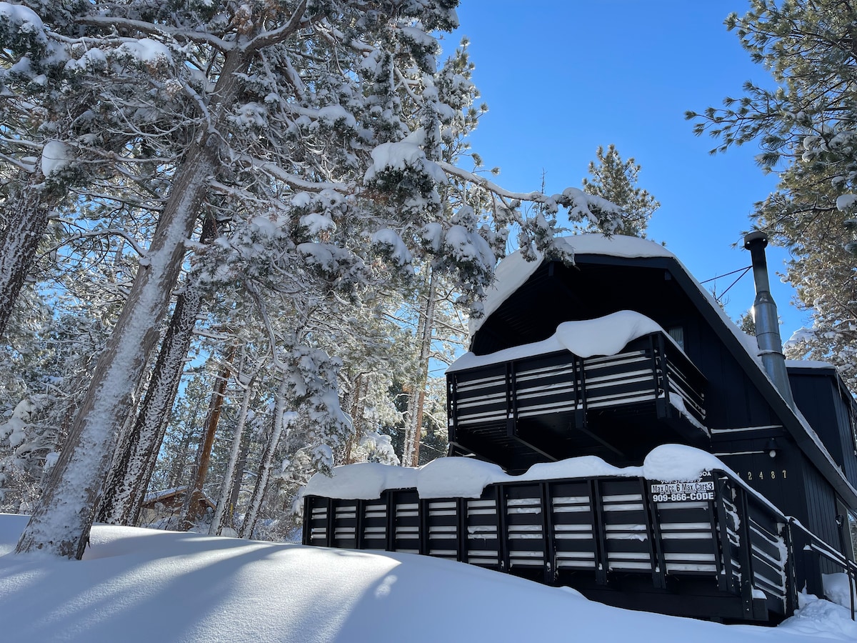 A black cabin is surrounded by tall snow-covered trees under a clear blue sky. Snow blankets the deck and roof, while the structure features distinct layered balconies, presenting a cozy atmosphere in the winter landscape.