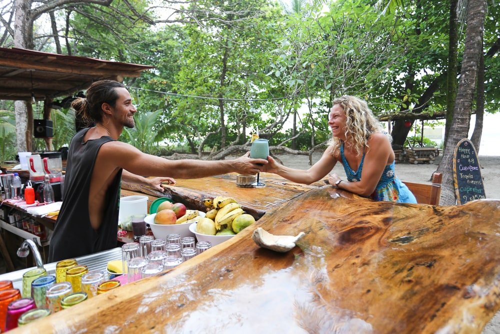 A rustic bar area features a large wooden counter that displays various colorful drinks. Lush greenery surrounds the setting, providing a natural backdrop. A bartender hands a drink to a patron, who is seated in a vibrant setting, enhancing the relaxing beach vibe.