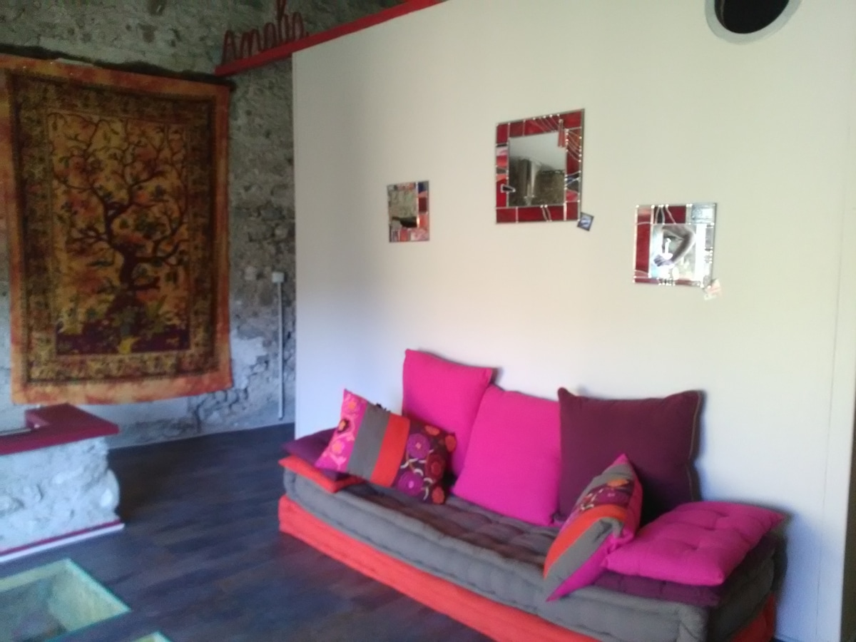 A vibrant seating area features a long cushioned sofa adorned with colorful pillows. Brightly colored wall decorations, including mirrors, complement a large, ornate tapestry hung on the stone wall, creating a distinct and warm living space.