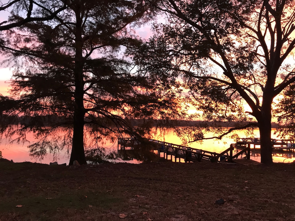 A serene lakeside scene features a private dock framed by large trees. The sky is painted with shades of orange and pink during sunset, reflecting softly on the water surface, creating a tranquil atmosphere for relaxation.