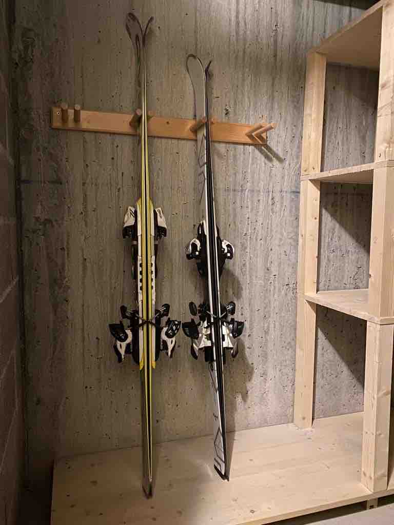 Two pairs of skis are mounted on a wooden rack against a concrete wall. A wooden shelf unit stands nearby, offering additional storage space. The minimalist design emphasizes functionality, providing an organized area for ski equipment.