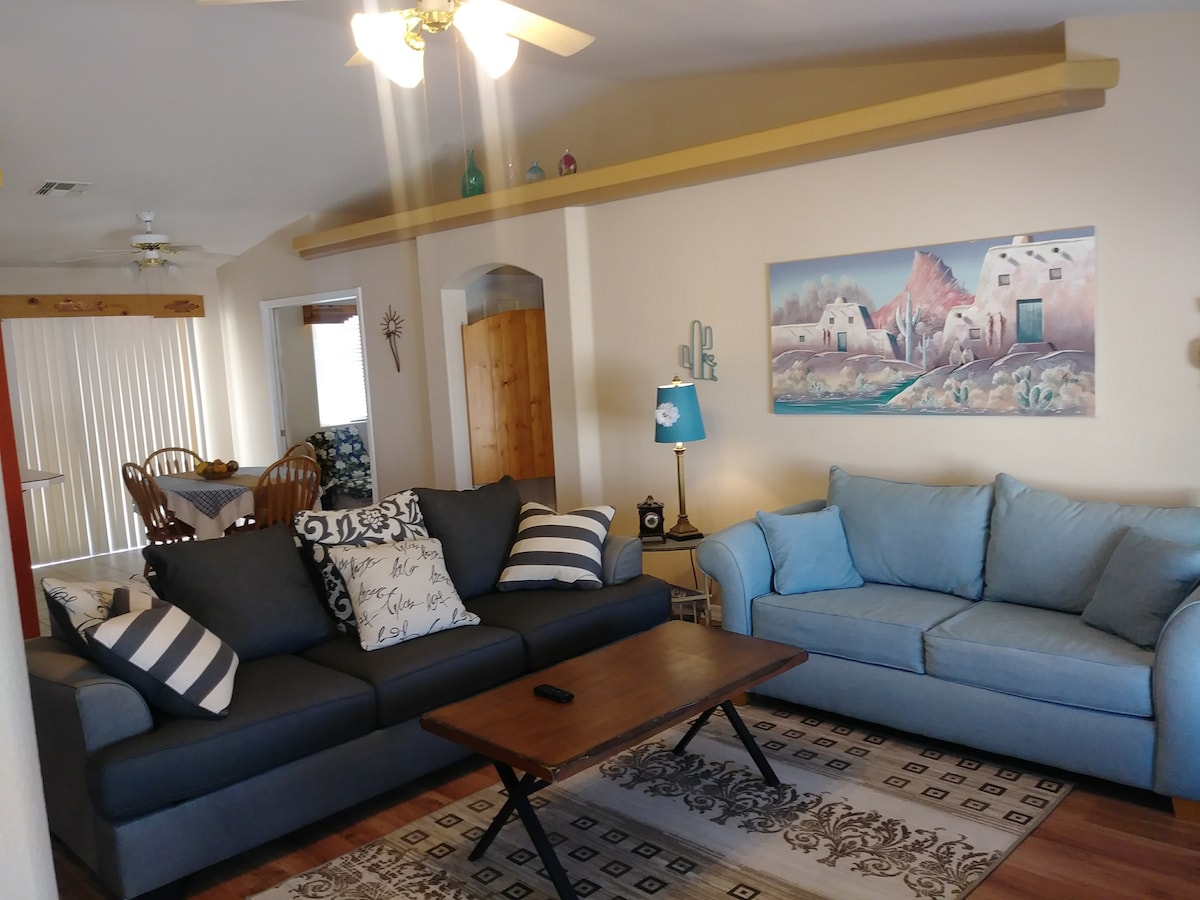 A bright living area features a mix of comfortable seating, including a dark sofa with patterned cushions and a light blue couch. A wooden coffee table takes center stage on a patterned rug, while artwork depicting a serene landscape is displayed on the wall.